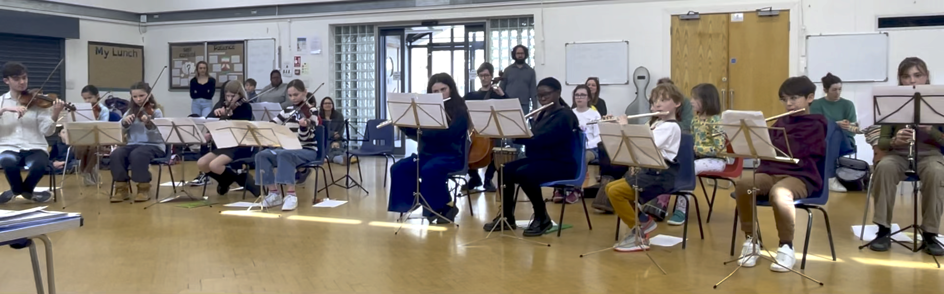 Members of the Junior Orchestra performing at the Spring 2024 concert. They are in a school hall. To the left there are students and staff seated and playing violins. To the right front, there are four students playing flute and one clarinet, and behind them students and staff playing cello and percussion. In the background there is a door to the playground with light coming in.